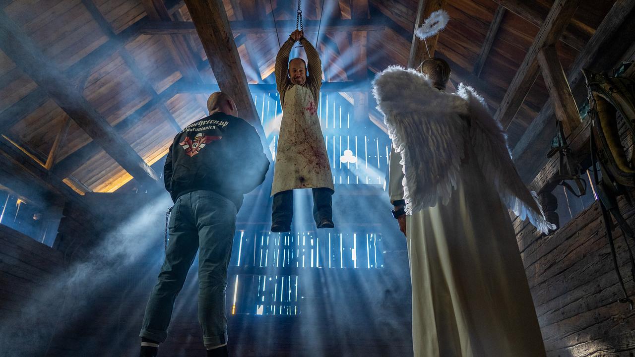 Roland Düringer hängt mit gefesselten Händen von der Decke eines hölzernen Dachbodens, während eine Person in Engelskostüm und ein Mann mit Rockerjacke am Boden stehen und zu ihm hinauf blicken.