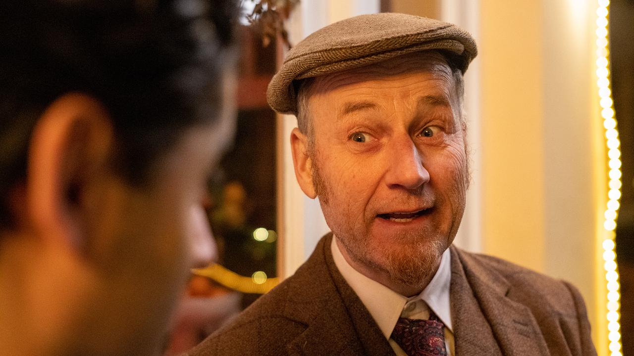 Roland Düringer mit Schirmmütze und braunem Tweedanzug steht in einem warm beleuchteten Innenraum und spricht mit jemandem.