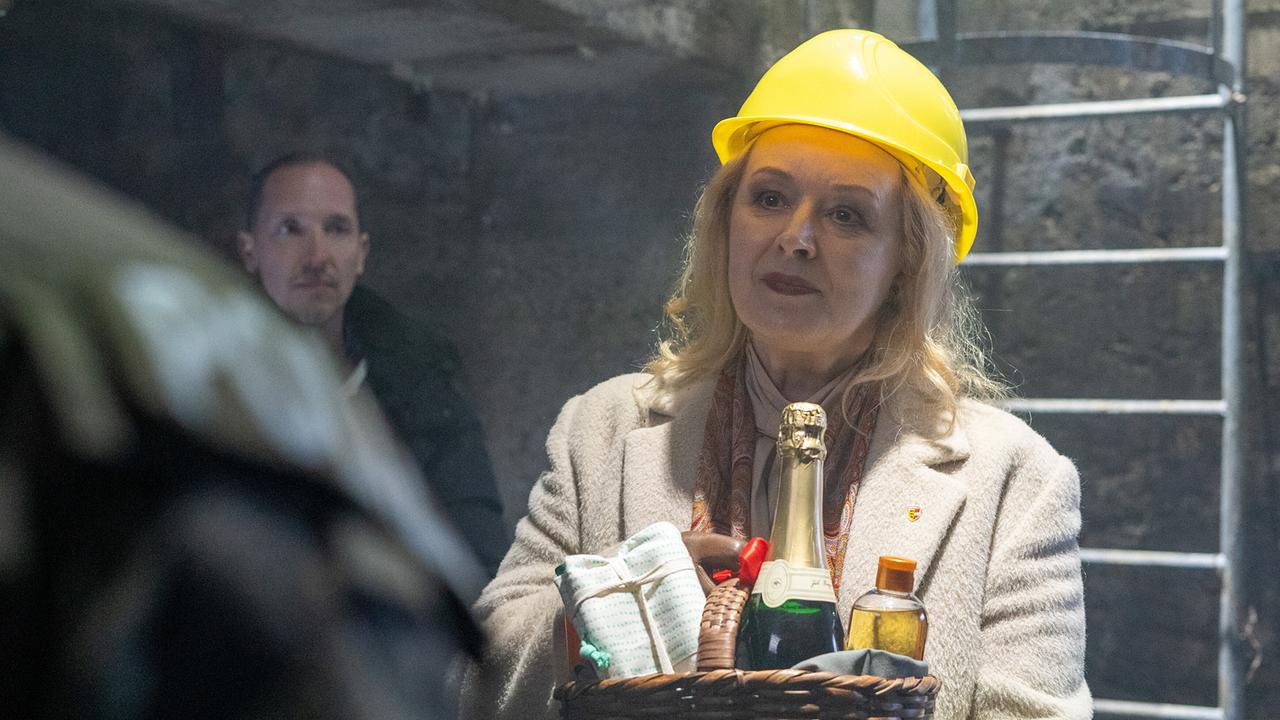 Petra Morzé mit gelbem Bauhelm hält einen Präsentkorb mit Sekt und Lebensmitteln in einem Schacht, um sie herum stehen zwei Männer.