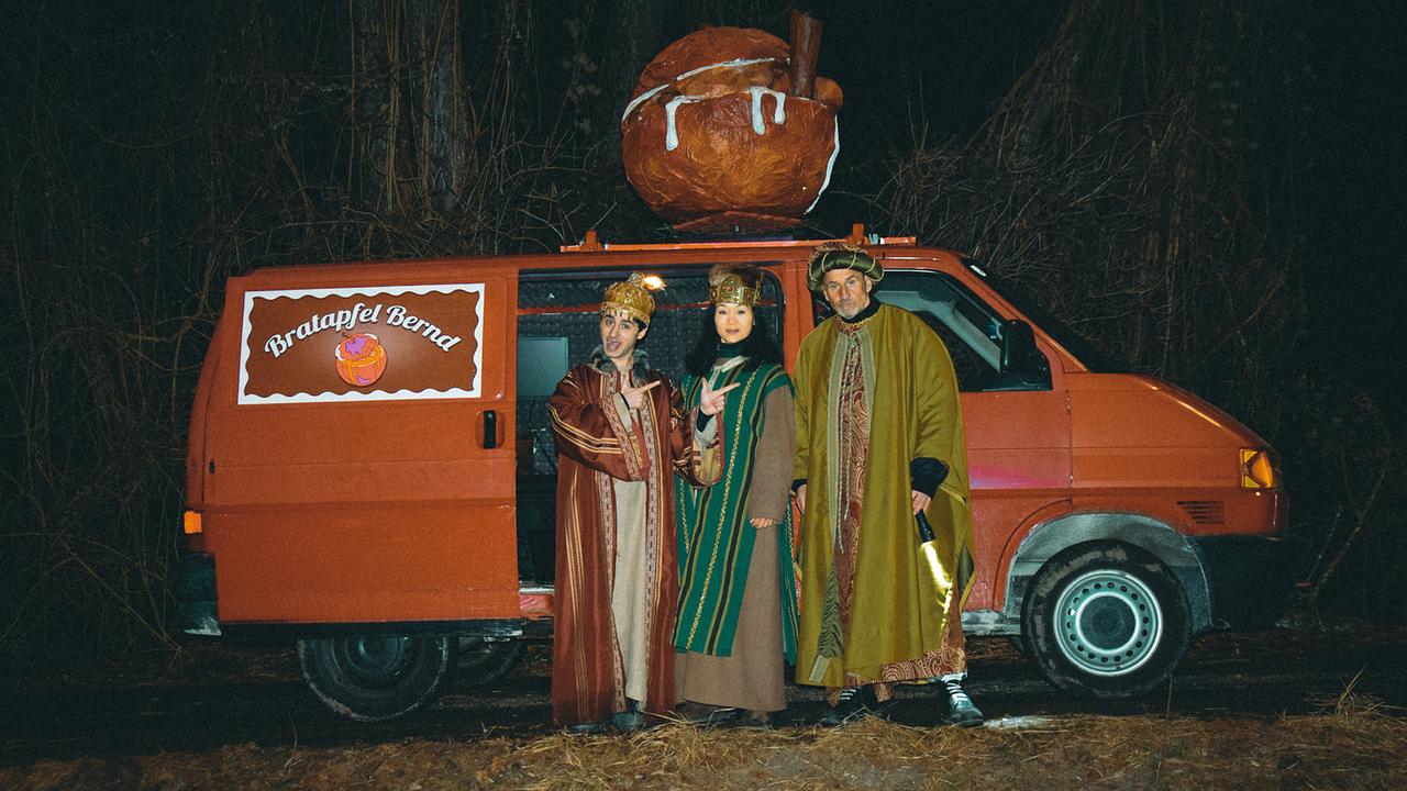 Enis, Domino und Kanakentöter Fred sind als Heilige Drei Könige verkleidet und stehen nachts vor einem roten Lieferwagen mit der Aufschrift „Bratapfel Bernd“ und einem großen Bratapfelmodell auf dem Dach.
