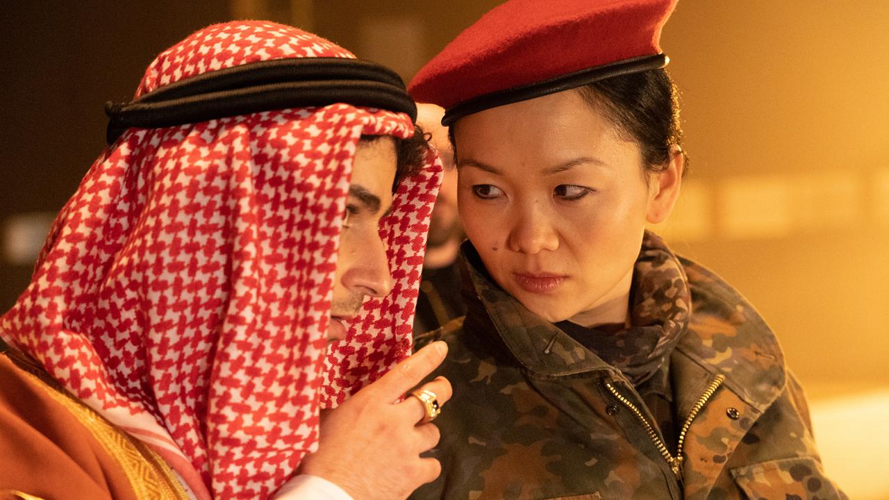 Enis in traditioneller arabischer Kleidung spricht eng mit Domino in Tarnuniform und rotem Barett.