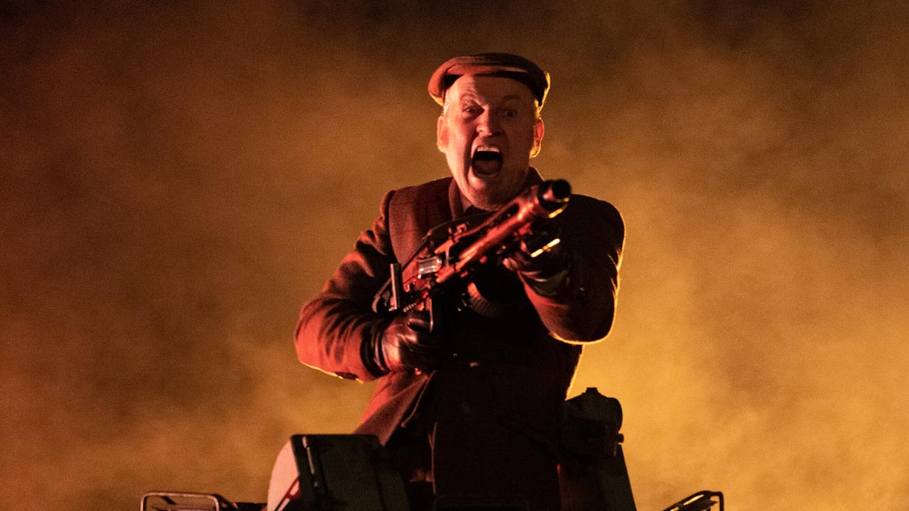 Roland Düringer in Militäruniform steht bewaffnet in einem Panzer vor einer Kulisse aus Rauch und orangem Licht.