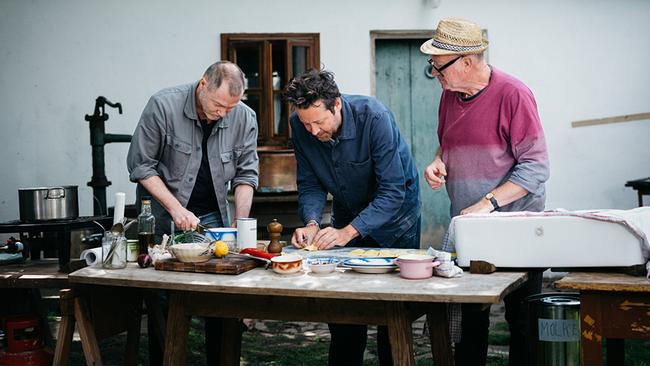 Florian Holzer, Ingo Pertramer und Thomas Nowak stehen an einem Außentisch und bereiten gemeinsam eine Mahlzeit zu, während verschiedenes Kochgeschirr und Zutaten vor ihnen ausgebreitet sind.