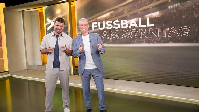 © ORF/Roman Zach-Kiesling Peter Stöger und Thomas Seidl stehen lächelnd im Studio von „Fußball am Sonntag“ mit Schriftzug an der Wand
