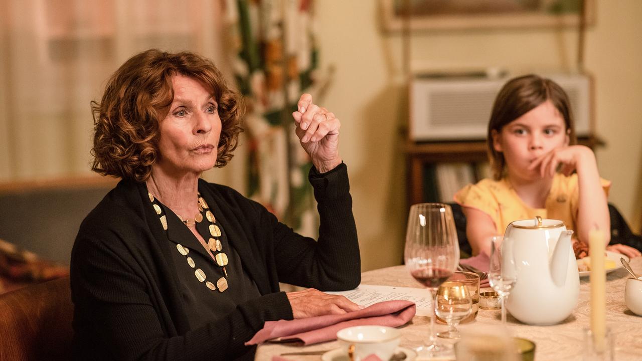 © ORF/PRO7/Warner Eine elegant schwarz gekleidete Frau und ein Kind in einem gelben Kleid sitzen gemeinsam an einem Wohnzimmertisch, auf dem Gläser, Tassen und Kannen stehen