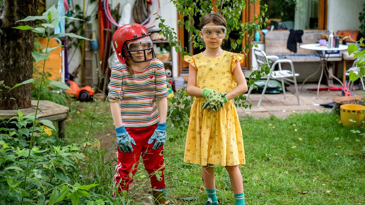 © ORF/PRO7/Warner Zwei Kinder stehen im Garten, eines mit rotem Football-Helm, bunt gestreiftem Shirt, das andere im gelben Kleid - beide tragen Gartenhandschuhe und Schutzbrillen