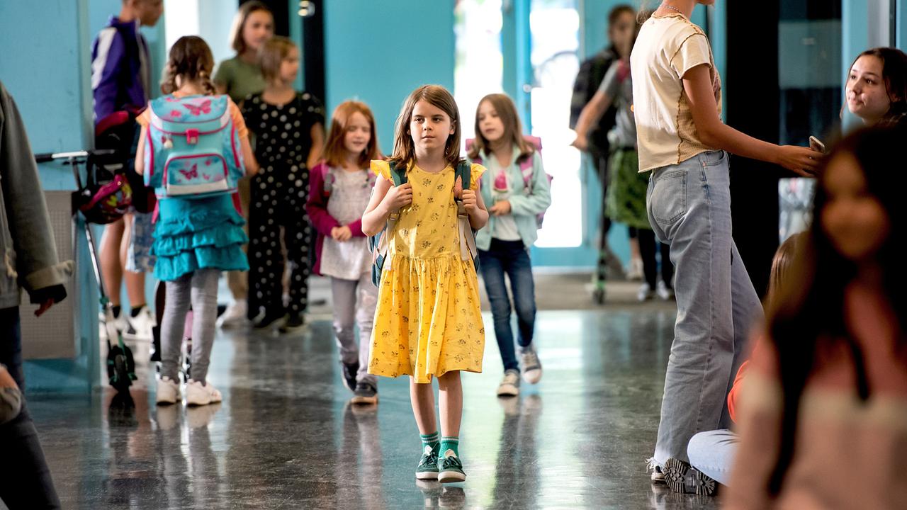 © ORF/PRO7/Warner Ein Schulkind in einem gelben Kleid und grünen Schuhen geht durch einen hellen Gang, umgeben von anderen Kindern mit bunten Schul-Rucksäcken