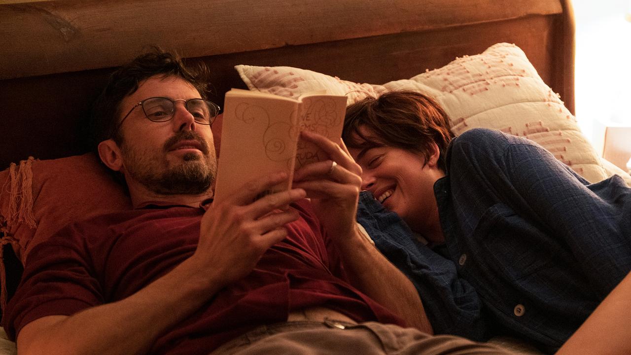 Ein Mann mit Brille und eine Frau liegen bekleidet nebeneinander im Bett, er liest ein Buch vor, sie lacht vergnügt