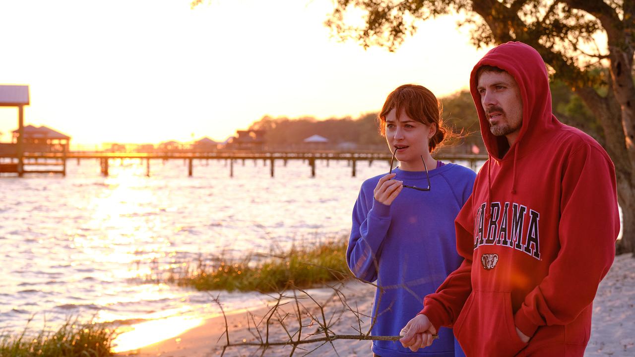 Ein junges Paar stehen am Ufer eines Sees, sie trägt einen blauen Sweater, er einen roten Hoodie mit der Aufschrift 'Alabama', hinter ihnen steht die Sonne tief am Horizont
