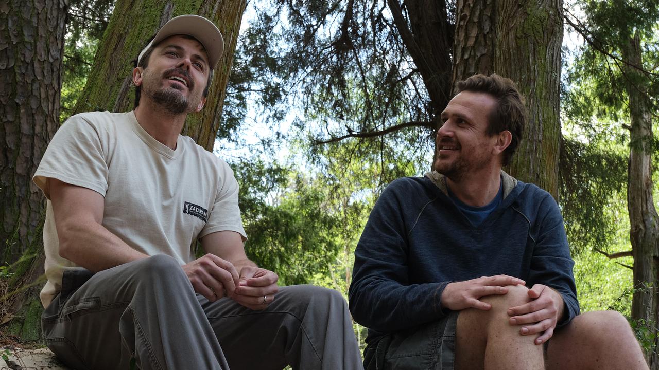 Zwei Männer machen bei einer sommerlichen Wanderung Rast unter schattigen Bäumen und unterhalten sich miteinander