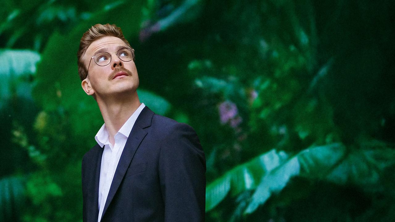 Matthias im Anzug und mit Brille blickt vor einem üppig grünen Hintergrund nach oben.