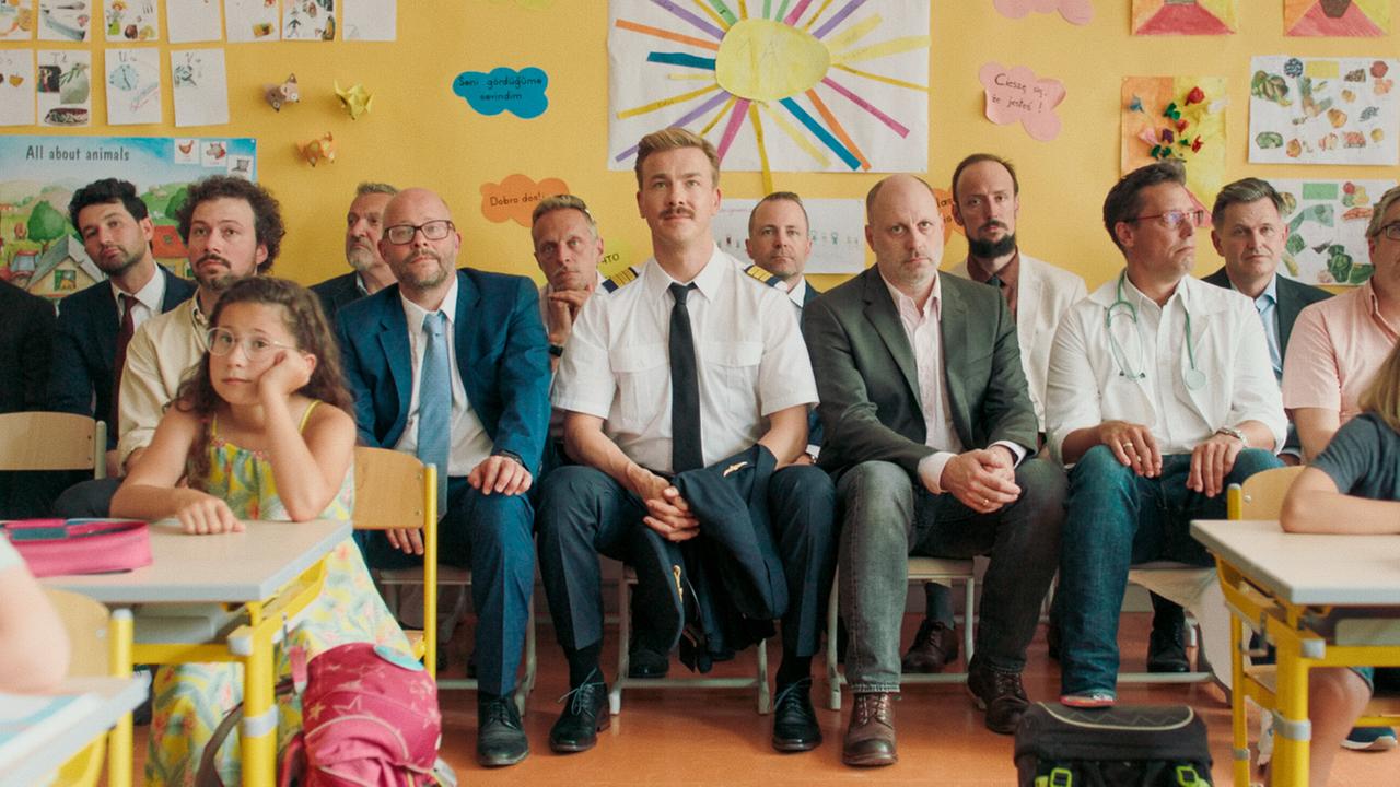 In einem Klassenzimmer sitzen zahlreiche Männer zwischen Schulkindern, darunter Matthias in Pilotenuniform.