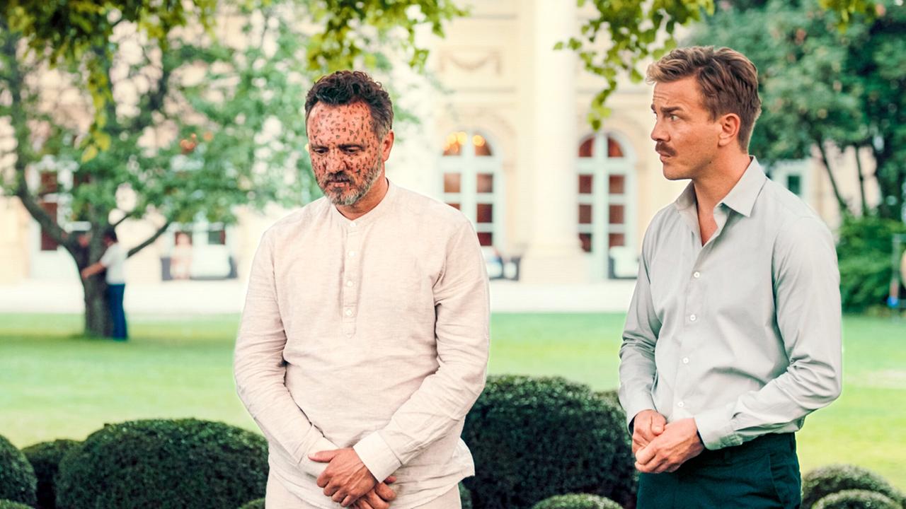 Matthias sucht Antworten im Yoga-Retreat und steht neben einem weiteren Mann im Garten eines eleganten Gebäudes.