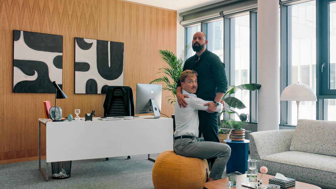 Matthias auf einem Sitzpolster umarmt seinen besten Freund und Kollegen David in einem modernen Büro.