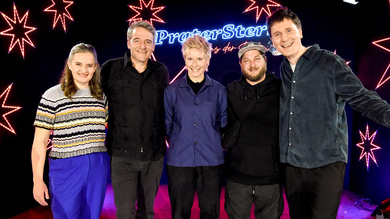© ORF/Hubert Mican Elli Bauer, Christoph Spörk, Anna Mabo, David Scheid und Hosea Ratschiller stehen gemeinsam vor einer bunt beleuchteten Bühne mit sternförmigen Lichtern und dem Schriftzug „PraterSterne“.