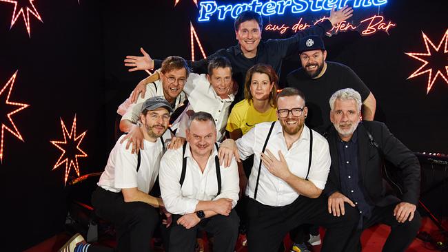 Hosea Ratschiller mit Band 5/8erl in Ehren, Dirk Stermann, Suse Lichtenberger und Michael Bauer stehen auf der Bühne mit leuchtenden Sternen und Neon-Schriftzug "Pratersterne - Rote Bar".