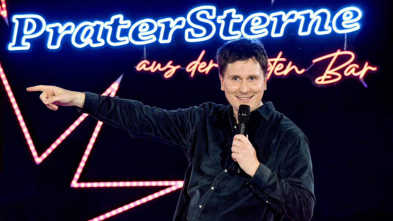 © ORF/Hubert Mican Hosea Ratschiller steht mit Mikrofon auf der Bühne mit leuchtenden Sternen und Neon-Schriftzug "Pratersterne - aus der Roten Bar".