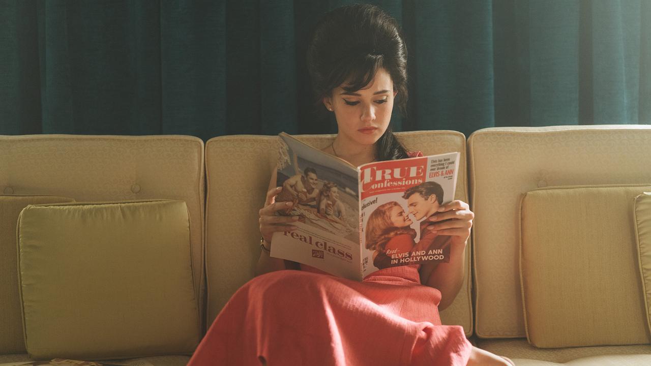 Eine junge Frau sitzt im rosaroten Kleid auf einer Couch und liest ein Magazin, auf dem Cover ist 'Elvis and Ann' zu lesen