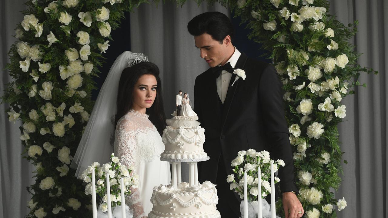 Ein Hochzeitspaar vor einem Torbogen weißer Rosen, die Braut weiß mit Krönchen und Schleier, der Bräutigam in schwarzem Anzug und weißer Rose am Revers - vor ihnen eine weiße Hochzeitstorte