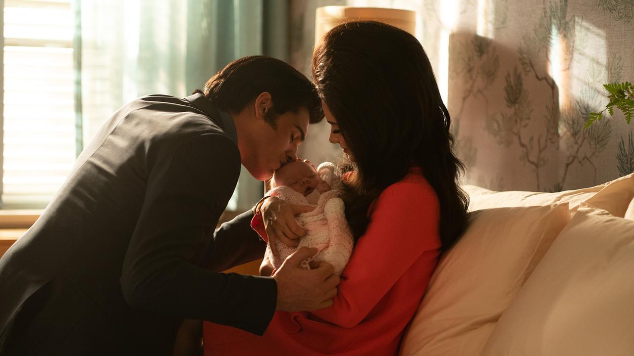 Eine Frau sitzt im rosaroten Schlafmantel auf dem Bett und hält ein Baby im Arm, ein schwarz gekleideter Mann beugt sich zu ihr und küsst das baby am Kopf