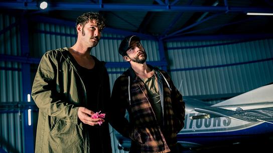 © ORF/Samsara Film Flo mit einer pinken Blume in der Hand und Eddi stehen nebeneinander in einem Hangar neben einem Kleinflugzeug.