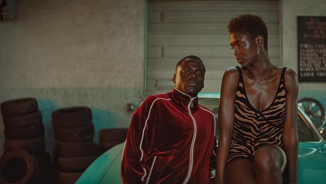 Retro-Atmosphäre - Slim in rotem Trainingsanzug und Queen in einem Kleid in Tiger-Look vor einem türkisfarbenen Oldtimer der 1970er Jahren in einer Werkstatt, umgeben von Reifen und Felgen