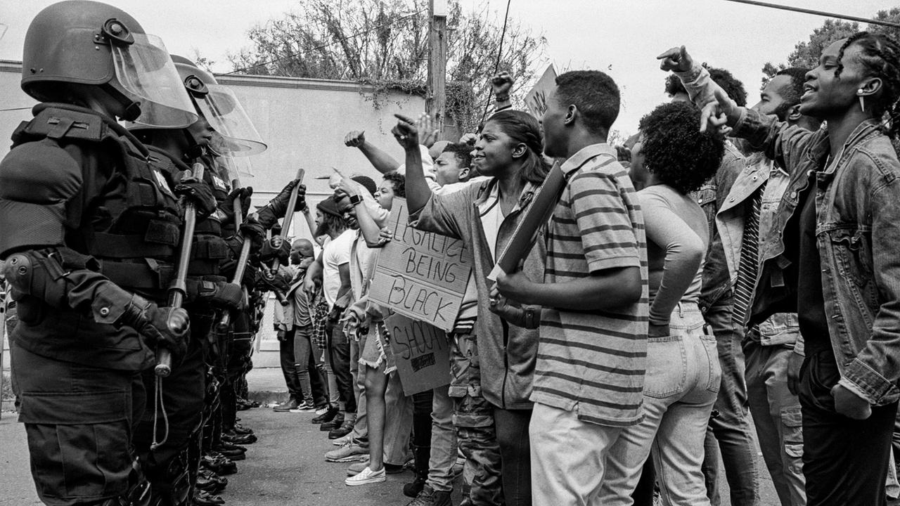 © ORF/Universal Pictures/Andre D. Wagner Demonstrierende stehen Polizisten in voller Schutzmontur gegenüber, ein Schild trägt die Aufschrift "LEGALIZE BEING BLACK"