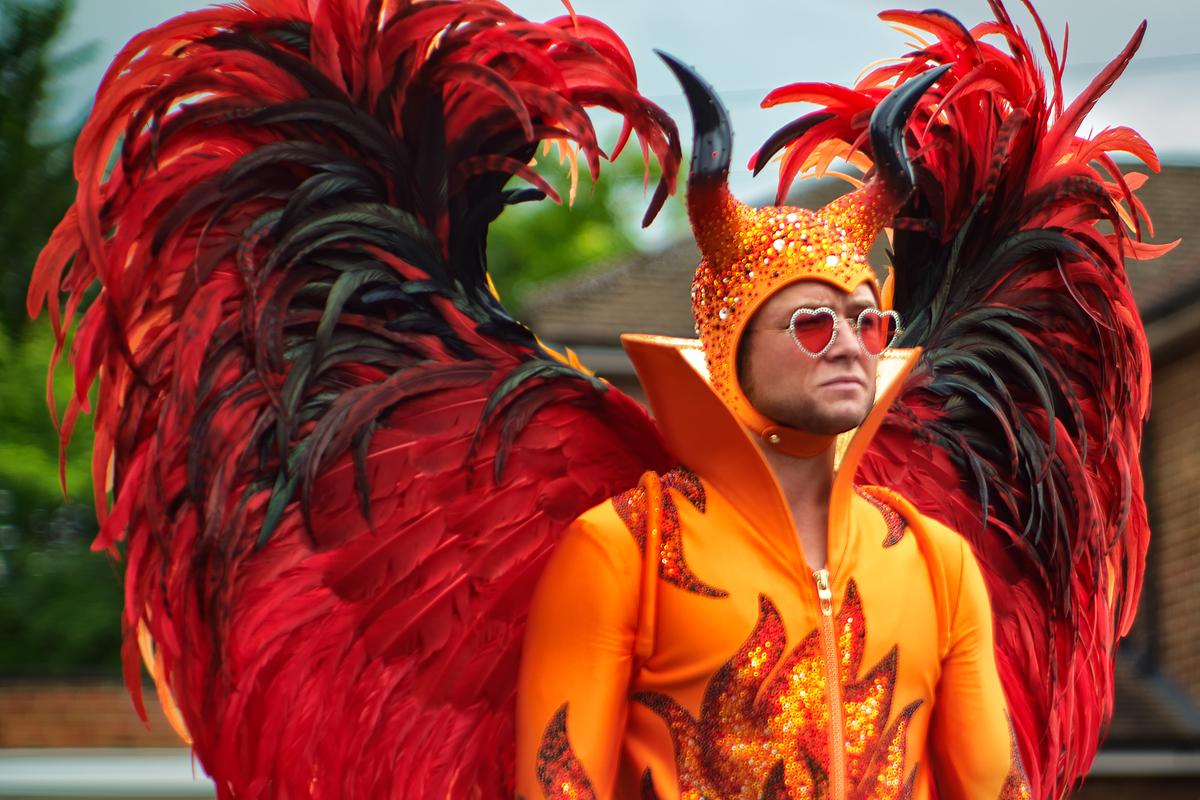 Taron Egerton als Elton John in einem feuerroten und orangefarbenden Bühnen-Outfit mit rot-schwarz gefiederten Flügeln und geschwungenen Hörnern auf dem Kopf