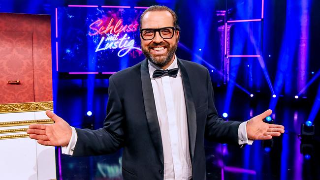 © ORF/Hubert Mican Gerald Fleischhacker im Smoking und Mascherl steht auf der bunt ausgeleuchteten "Schluss mit lustig"-Showbühne und gestikuliert offen in Richtung Publikum.