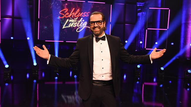 Gerald Fleischhacker im Smoking und Mascherl steht auf der bunt ausgeleuchteten "Schluss mit lustig"-Showbühne und gestikuliert offen in Richtung Publikum.