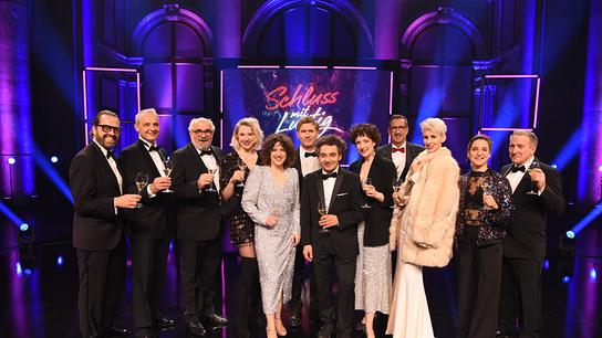 Gerald Fleischhacker, Thomas Maurer, Michael Niavarani,    Jennifer Frankl, Ariana Schirasi-Fard, Joachim Brandl, Matthias Mamedof, Katharina Dorian, Viktor Gernot, Lisa Eckhart, Christina Kiesler und Robert Palfrader, alle festlich gekleidet, posieren auf einer Bühne mit der Aufschrift „Schluss mit Lustig“ mit Sektgläsern.