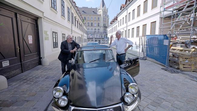 Michael Niavarani und  Harald Schmidt stehen neben einem schwarzen Oldtimer auf einer Kopfsteinpflaster-Straße in Wien