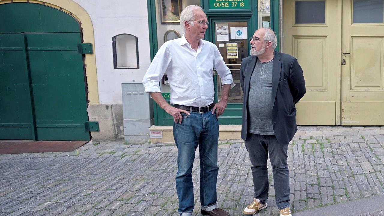 © ORF/Peter Koköfner Harald Schmidt und Michael Niavarani stehen beim Stadtspaziergang auf einer gepflasterten Gasse vor alten Gebäuden und unterhalten sich.