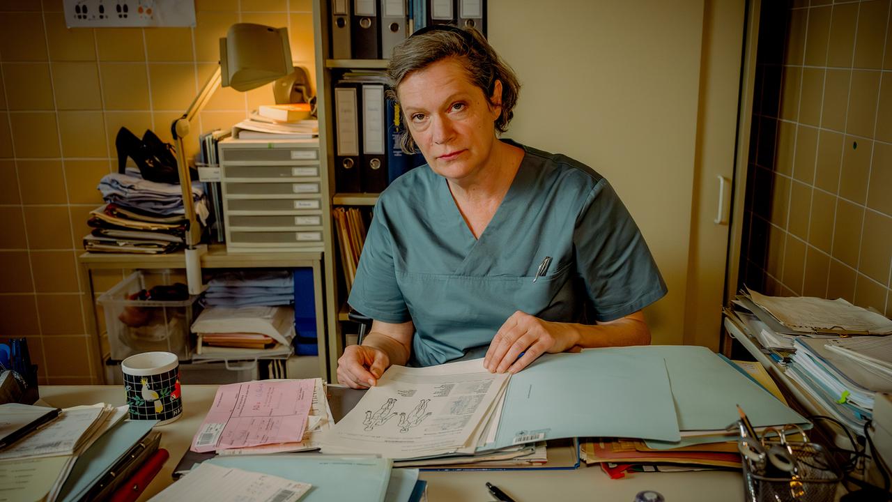 Elisabeth Imler in medizinischer Arbeitskleidung sitzt an einem Schreibtisch voller Akten und Unterlagen.