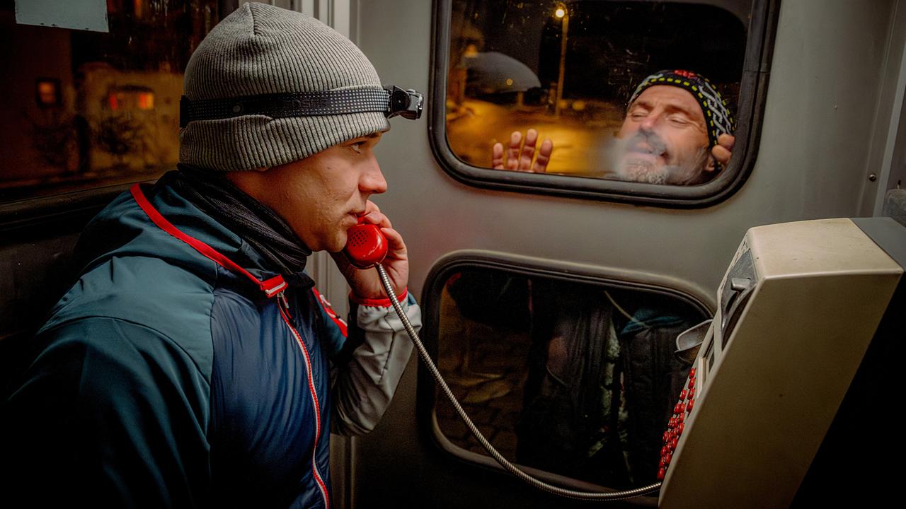 Martin Steiner telefoniert in einer alten Telefonzelle, während draußen Egbert durch die Glastür schaut.