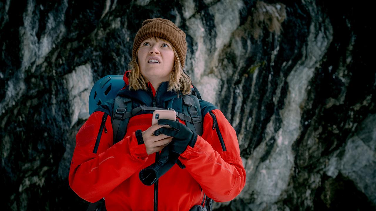Melanie Grandits  in roter Winter-Outdoor-Kleidung steht vor einer Felswand und hält ein Smartphone.