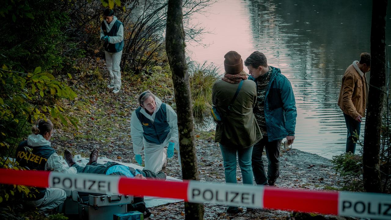 Ermittler arbeiten an einem abgesperrten Tatort am Seeufer, während Melanie Grandits und Martin Steiner miteinander sprechen.