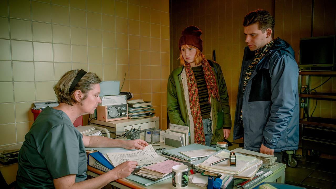 Marlene Hauser und Simon Morzé  stehen in einem Büro vor dem Schreibtisch von Elisabeth Imler, die Unterlagen durchsieht.