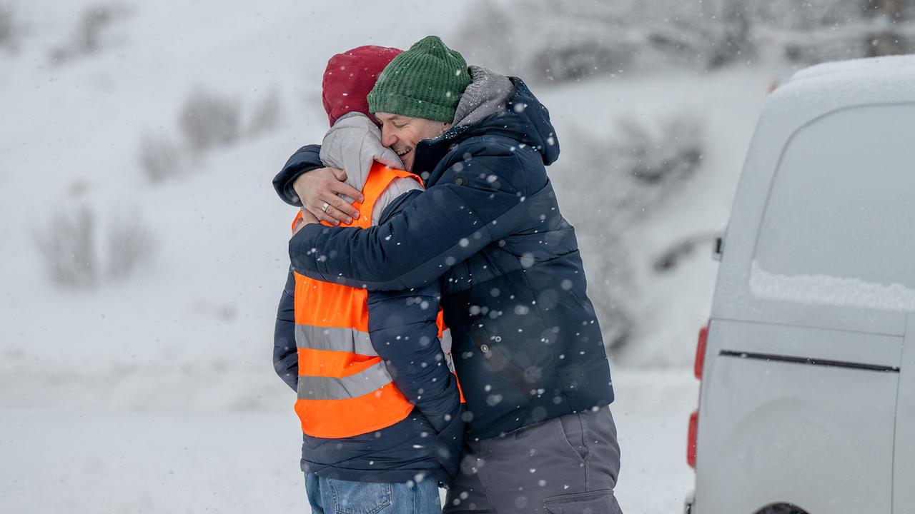 Mario Rossi umarmt Luca Rossi im Freien bei starkem Schneefall neben einem Fahrzeug.
