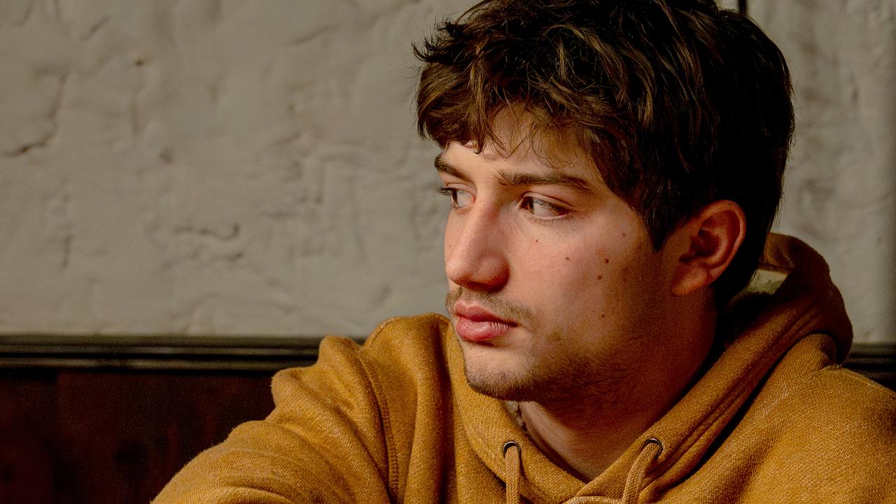 Nikki Auer sitzt mit ernstem Blick seitlich gedreht auf einer Holzbank.