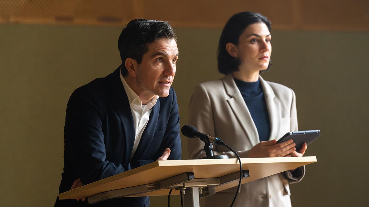 Tony steht an einem Rednerpult, daneben steht seine Assistentin