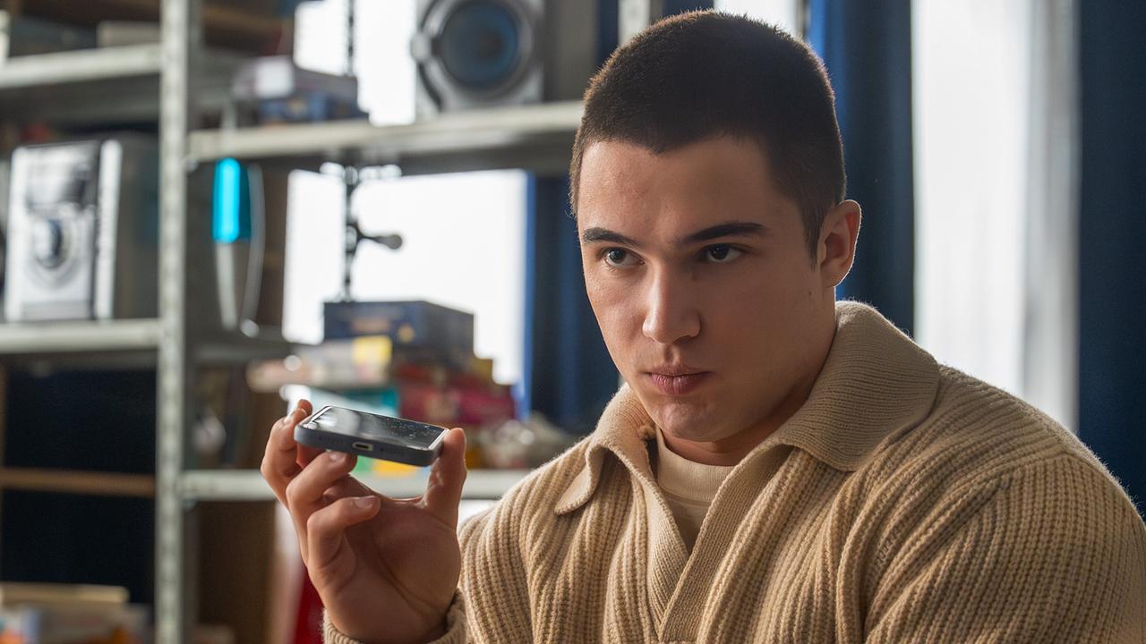Georg Stoiber  sitzt in einem Raum und hält ein Smartphone in der Hand, als würde er etwas abspielen.