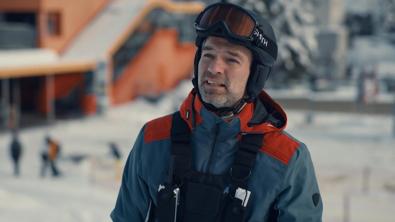 Mark Auer in Skikleidung trägt Helm und Skibrille vor einem verschneiten Skigebiet.