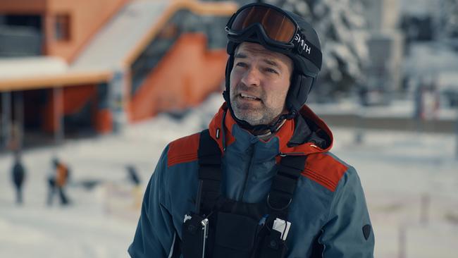 © ORF/BR/Superfilm Mark Auer in Skikleidung trägt Helm und Skibrille vor einem verschneiten Skigebiet.