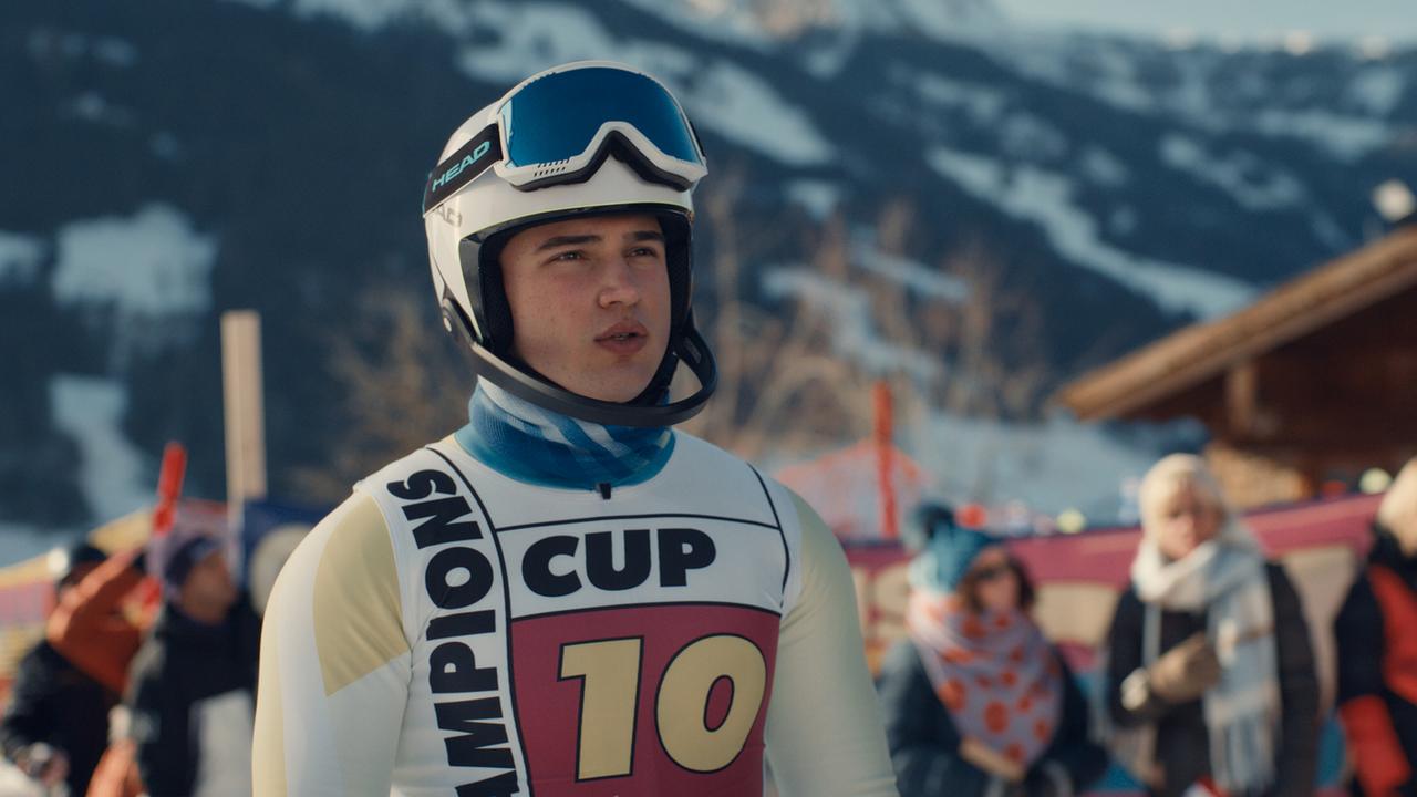 Georg Stoiber im Ski-Rennanzug und Helm steht im Publikumsbereich vor einer Bergkulisse.