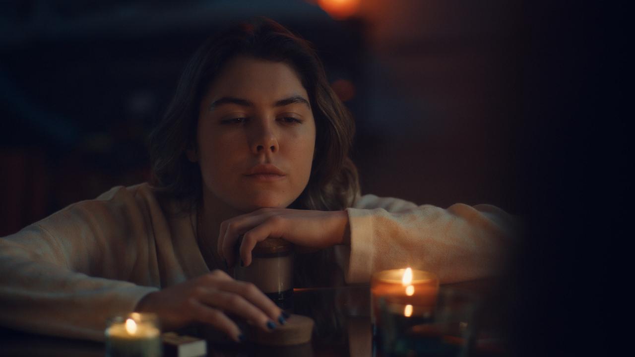 Solenn Eden sitzt an einer Theke in einem dunklen, stimmungsvoll beleuchteten Raum und stützt ihr Gesicht auf einen Kaffeebecher, umgeben von brennenden Kerzen.