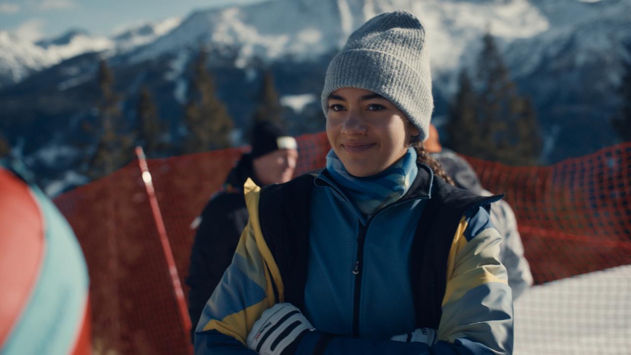 Nawal Ammann in Winterkleidung und mit Haube steht bei Sonnenschein vor einem roten Sicherheitsnetz in einer verschneiten Berglandschaft.