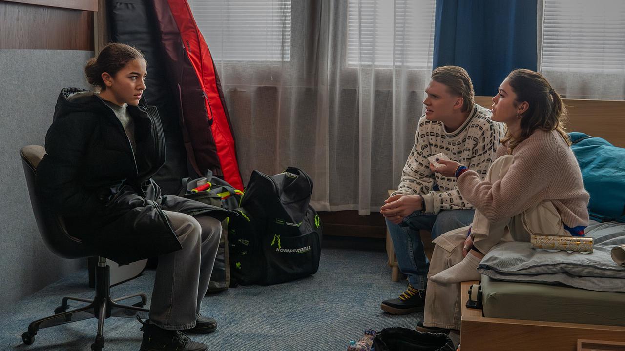 Nawal Ammann sitzt auf einem Drehsessel in einem Zimmer Bobby Harrer und Solenn Eden gegenüber, die auf einem Bett sitzen.