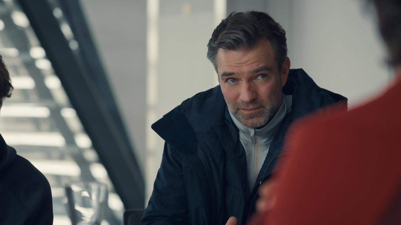 Mark Auer in Winterkleidung spricht in eiem modernen Raum mit Personen ihm gegenüber.