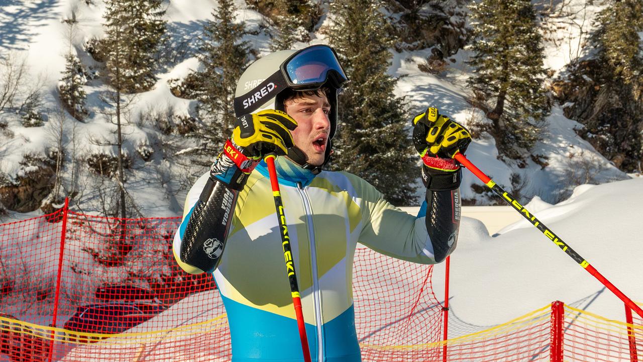Nikki Auer im Skianzug steht mit Skistöcken vor einem Sicherheitsnetz und schneebedecktem Gelände.